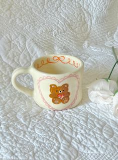 a teddy bear mug sitting on top of a bed next to a white flower vase