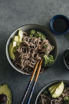 two bowls filled with noodles, broccoli and avocado next to chopsticks