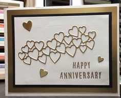 a happy anniversary card with gold hearts on white paper and black border, in front of bookshelves