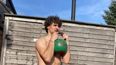 a shirtless man holding a green object in front of a wooden wall and fence