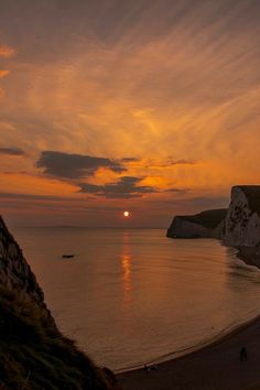 the sun is setting over the ocean with people walking on the beach and cliffs in the background