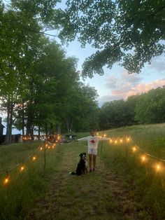 a man standing next to a black dog on top of a grass covered field with lights strung across it