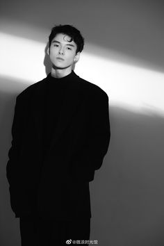 a man standing in front of a white wall wearing a black suit and turtle neck sweater