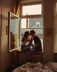 two people sitting on a window sill in front of a bed and looking out the window