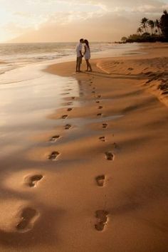 a man and woman standing on top of a beach next to the ocean with footprints in the sand