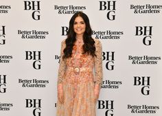 a woman standing in front of a white wall wearing a long dress and smiling at the camera