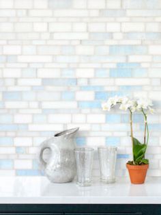 a white vase with flowers and two glasses on a table next to a brick wall