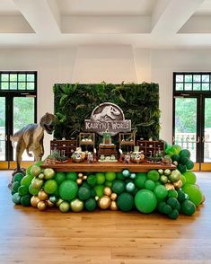 a table topped with green and gold balloons next to an artificial dinosaur statue on top of a hard wood floor