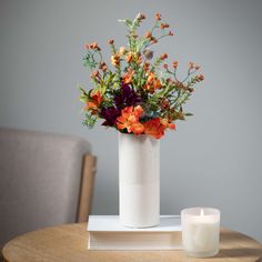 an arrangement of flowers in a white vase on a table with a lit candle next to it