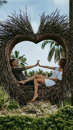 a man and woman sitting in a heart - shaped chair made out of branches with palm trees behind them