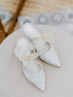 two white shoes with bows on them sitting on a cushion in front of a chair