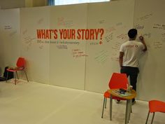 a man writing on a wall with chairs and tables in front of it that says what's your story?