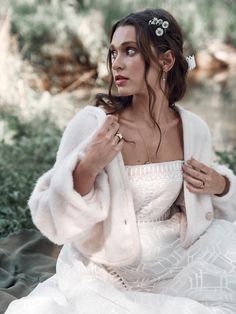 a woman sitting on the ground wearing a white dress and fur coat with flowers in her hair