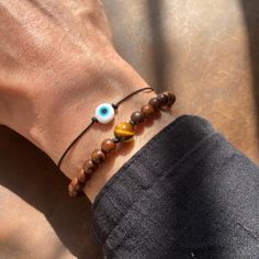 This bracelet set features two unique designs that complement each other perfectly. The first bracelet showcases a simple yet striking Evil Eye charm on a black adjustable cord, providing a touch of minimalistic style. The second bracelet is adorned with a single Tiger's Eye bead at its center, surrounded by rich, earthy wooden beads. Both bracelets are adjustable, making them suitable for various wrist sizes. This set is ideal for those who appreciate a blend of boho and earthy fashion styles, making it a versatile addition to any jewelry collection, whether for men or women. The Evil Eye is a powerful symbol of protection, believed to ward off negative energy and bring good fortune to the wearer. Its vibrant blue color is a timeless representation of safeguarding and positive vibes. The Spiritual Brown Bracelets For Friendship, Spiritual Brown Friendship Bracelets, Adjustable Spiritual Bracelet With Wooden Beads, Brown Round Beads Jewelry For Friendship, Adjustable Brown Wristband With 8mm Beads, Minimalist Brown Beaded Bracelets For Gifts, Minimalist Handmade Brown Bracelets, Adjustable Symbolic Bracelets With Round Beads, Adjustable Symbolic Bracelet With Round Beads