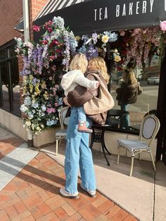 a woman holding a child in her arms while standing on the side walk next to a flower shop