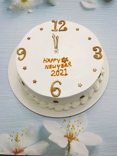 a white cake with gold numbers on it and flowers around the clock is sitting on a table