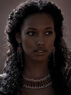 a woman with long dark hair wearing red earrings and necklaces on her neck, looking at the camera