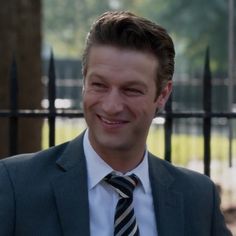 a man in a suit and tie smiles at the camera while standing next to a fence