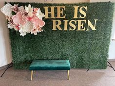 a green bench sitting in front of a fake grass wall with flowers and the words he is risen on it