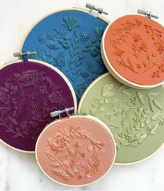 four embroidery hoops with flowers on them sitting on a marble surface, each one being embroidered in different colors