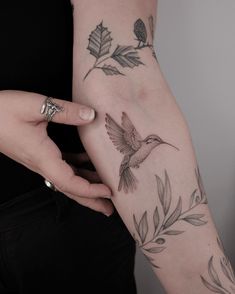 a woman's arm with tattoos on it and a hummingbird flying over leaves