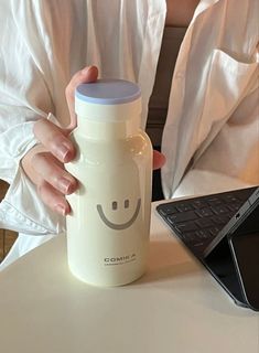 a woman is holding a water bottle with a smiley face on it and her laptop in the background