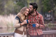 a man and woman standing next to each other on a wooden bridge holding coffee cups