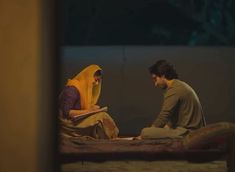 two people sitting on a bed looking at something in front of them with a book open