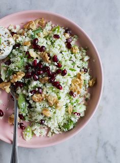 a pink bowl filled with rice, nuts and cranberries next to a spoon