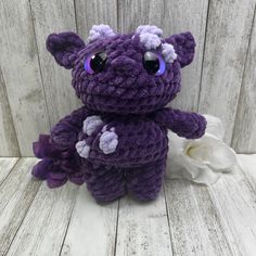 a purple crocheted teddy bear with big eyes and flowers on the bottom, sitting in front of a wooden wall