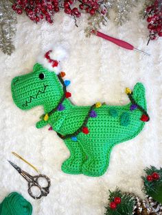 a green crocheted dinosaur laying on top of a white rug next to christmas decorations