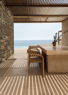 a wooden table sitting under a roof next to a stone wall near the ocean on a sunny day