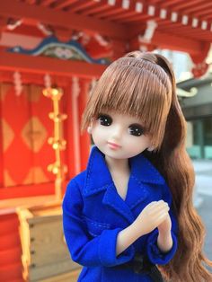 a doll with long hair standing in front of a red building wearing a blue coat