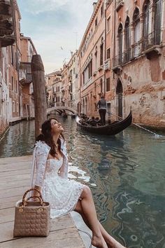 a woman sitting on the edge of a dock next to a canal with gondola boats in the background