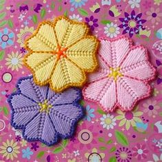 three crocheted flowers sitting on top of a pink and purple table cloth covered surface