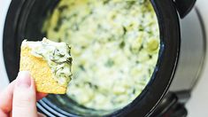 a hand holding a piece of bread over a crock pot filled with spinach dip