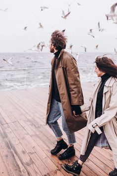 a man and woman walking on a pier with seagulls in the sky behind them