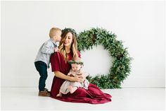 a woman holding a baby while standing next to a christmas wreath with a little boy on her lap