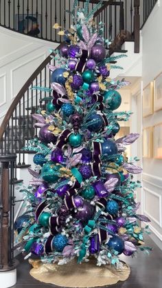 a christmas tree decorated with purple, green and blue ornaments in front of a spiral staircase