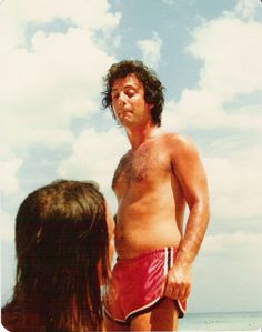 a shirtless man standing next to a woman on the beach looking at something in the sky