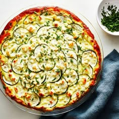 a pizza topped with zucchini and cheese on top of a white table next to a bowl of parsley