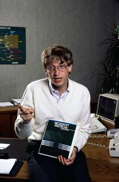 a man sitting at a desk with a book in his hand and a computer on the table behind him