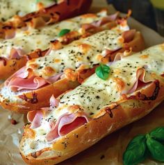 two hot dogs covered in cheese and ham on buns with basil leaves next to them