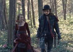 a man and woman dressed in period costumes walking through the woods with trees behind them