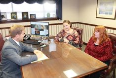 three people sitting at a table in front of a computer monitor and keyboard, talking to each other