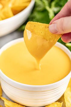 a hand holding a tortilla chip over a bowl of dip with chips in the background