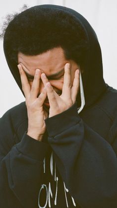 a man in a black hoodie covers his face with his hands as he stands against a white wall