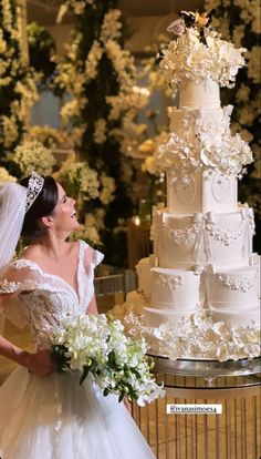 a woman in a wedding dress standing next to a tall white cake with flowers on it