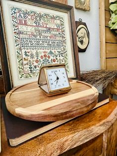 a clock sitting on top of a wooden table next to a framed cross stitch pattern