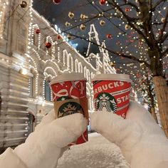 two cups of starbucks coffee are in the snow with christmas lights on the building behind them